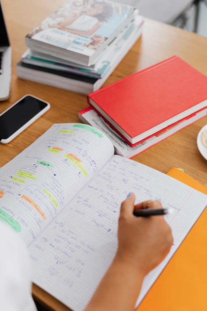 services-03 Student writing notes in a notebook during study session with books, phone, and highlighters.